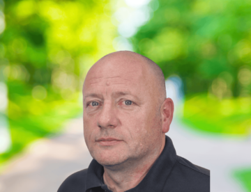 Man in a black polo shirt stood against a blurry backdrop with trees and a path.