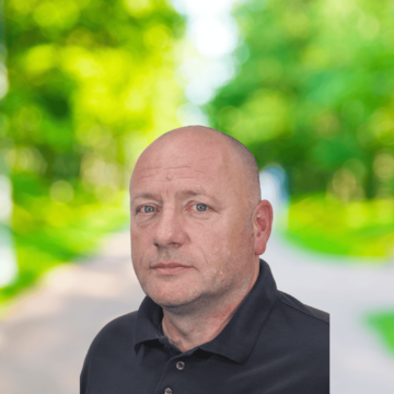 Man in a black polo shirt stood against a blurry backdrop with trees and a path.