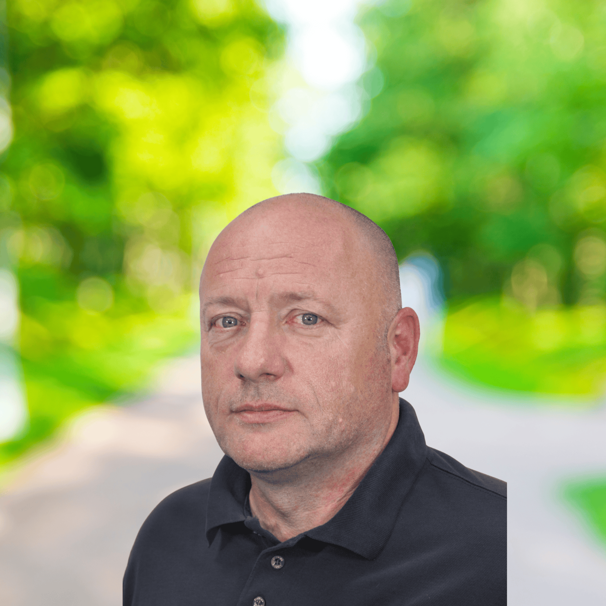 Man in a black polo shirt stood against a blurry backdrop with trees and a path.